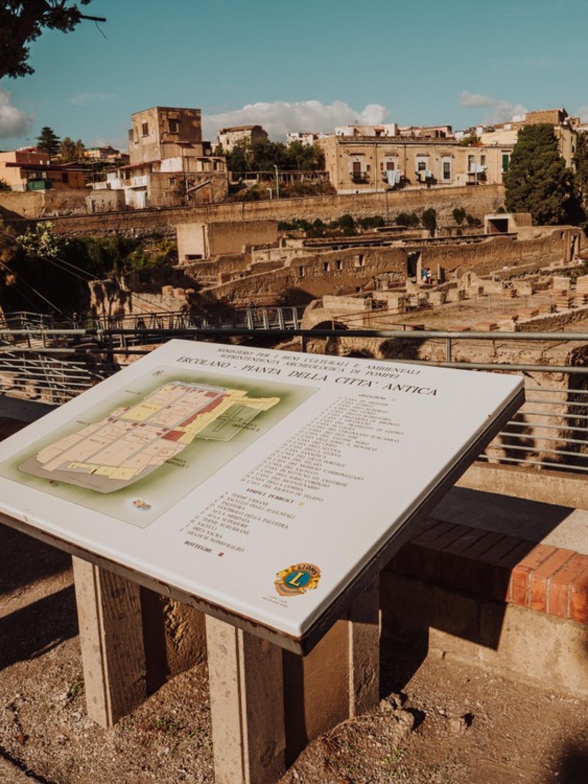 Information board at an ancient ruins site, with buildings and trees in the background.