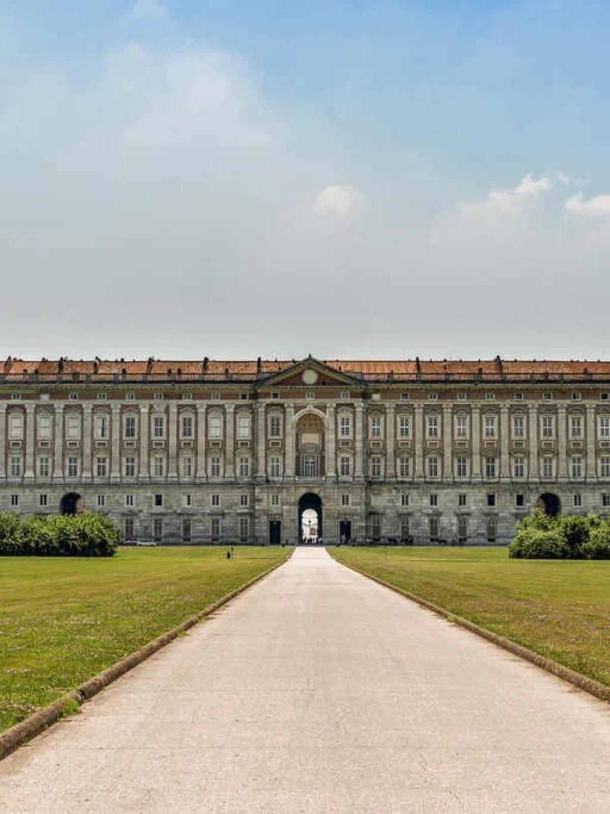 a castle with a clock at the top of a grass covered field with Palace of Caserta in the background