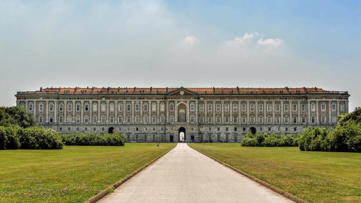 a castle with a clock at the top of a grass covered field with Palace of Caserta in the background