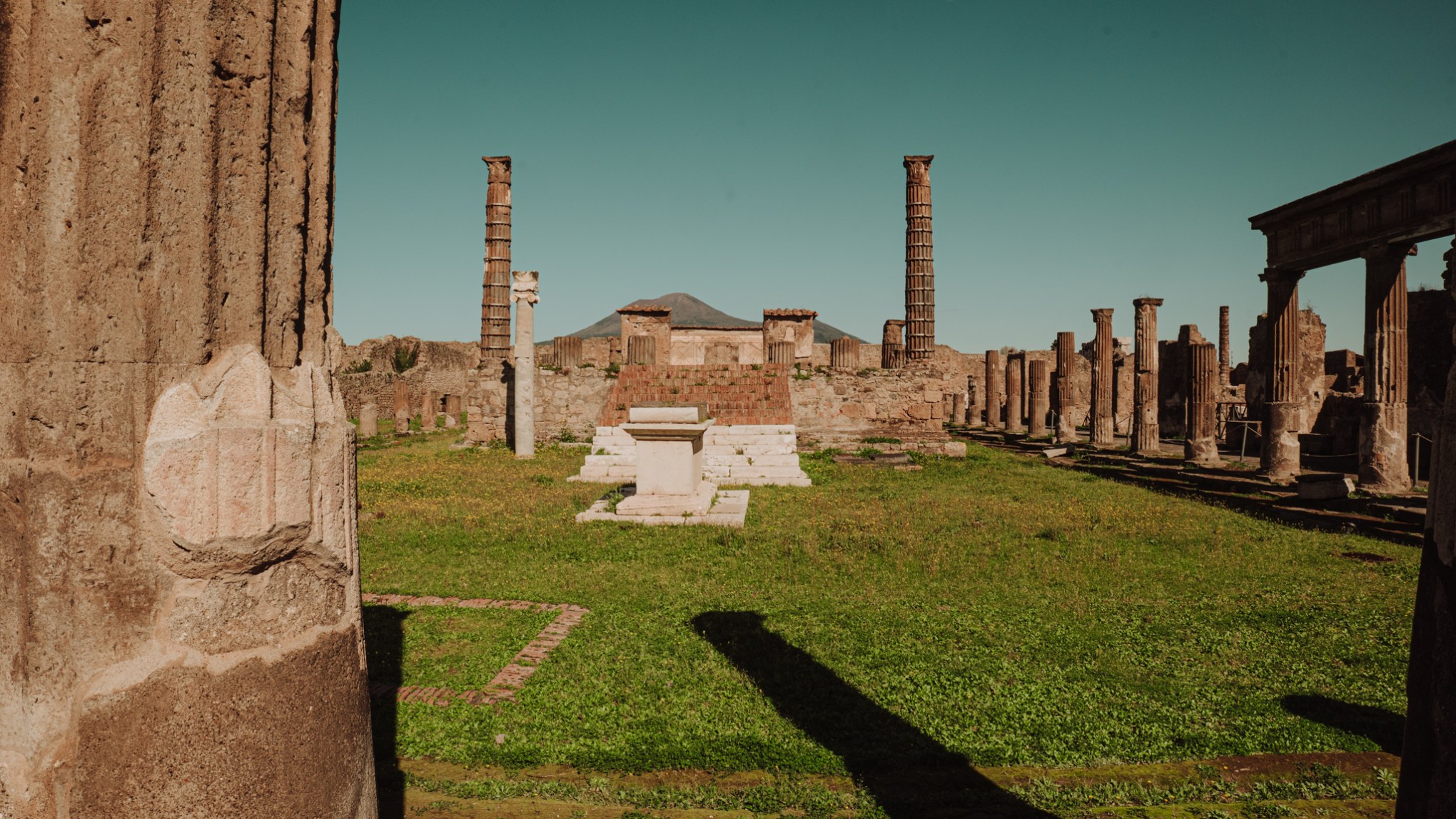 Ancient ruins with columns and a temple, grassy area, and mountain in the background.