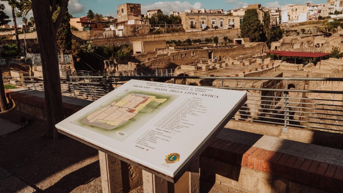 Information board in front of ancient ruins with buildings in the background on a sunny day.