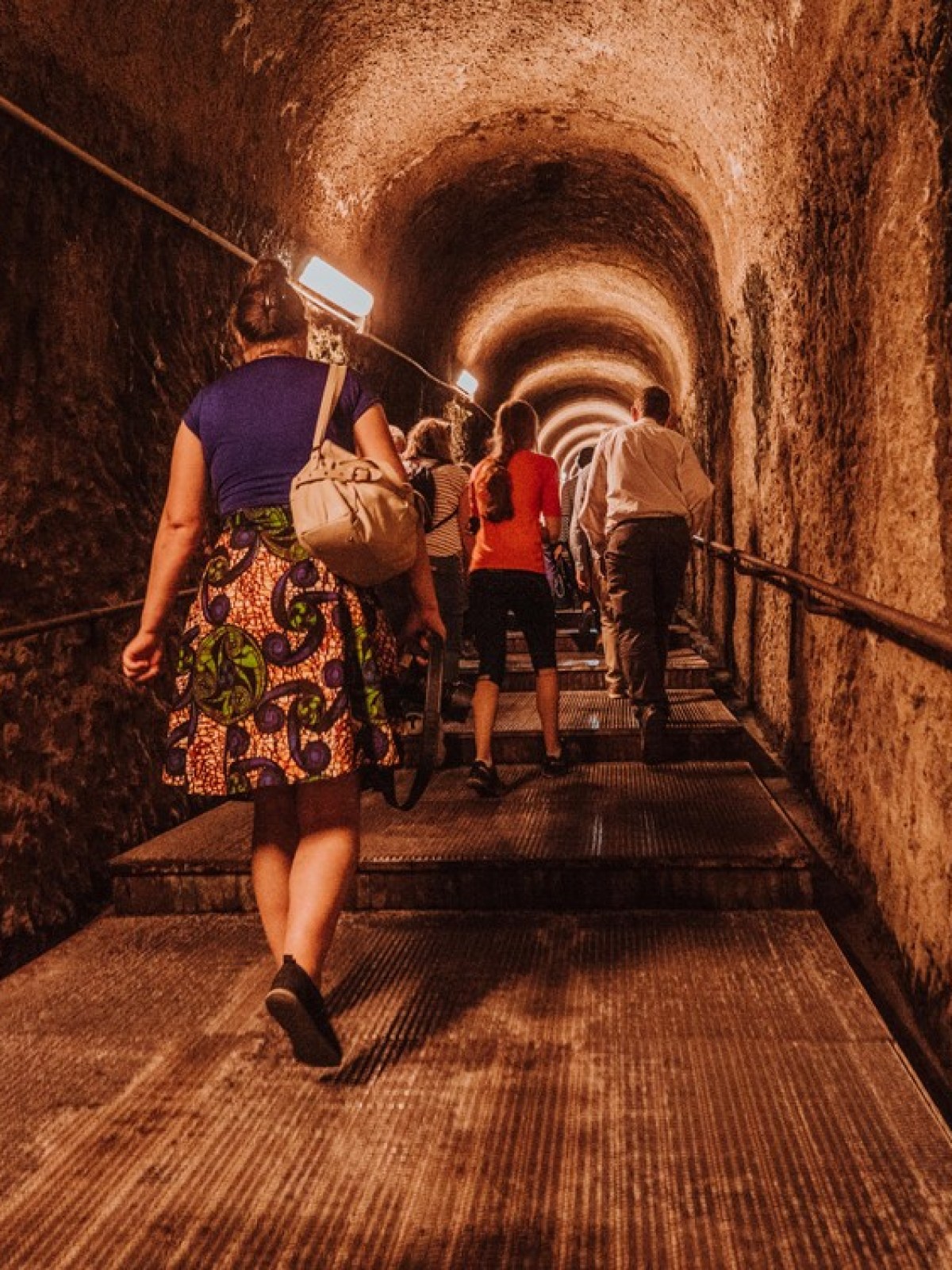 People walk up a dimly lit tunnel with stone walls and metal stairs.