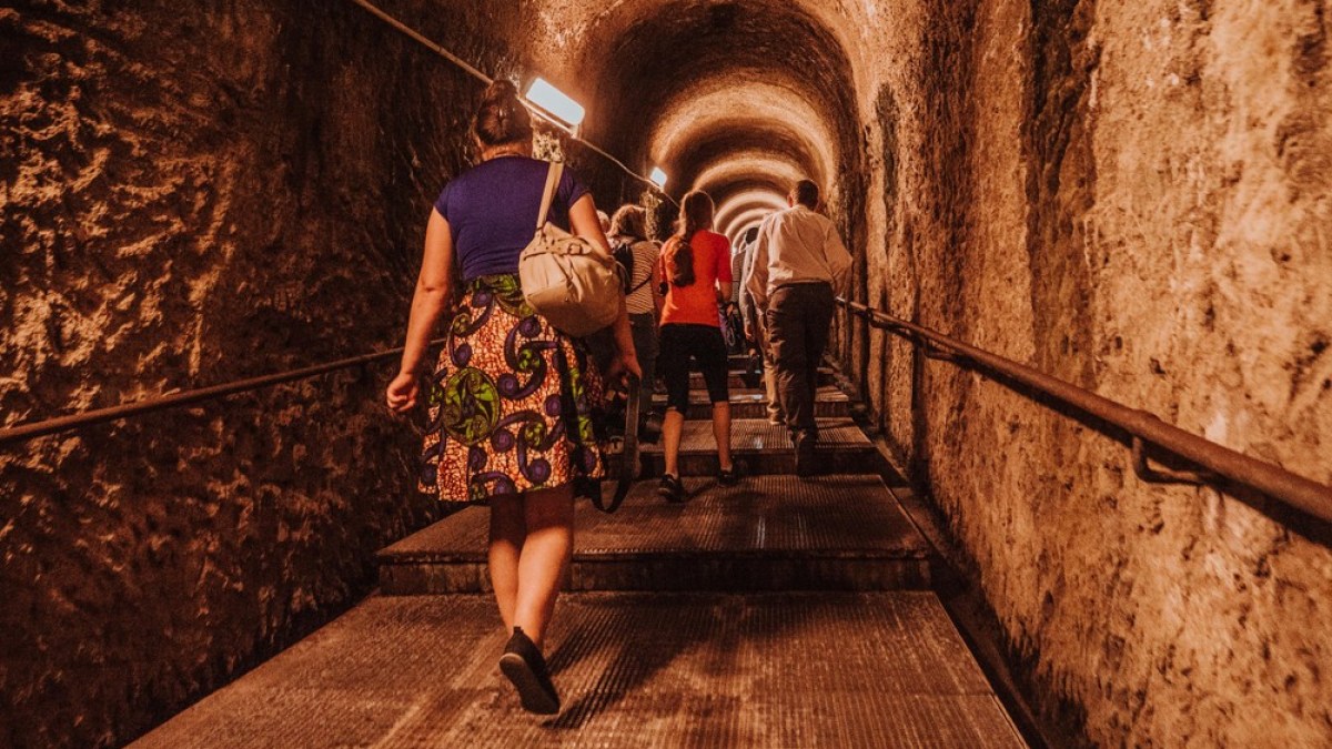 People walk up a dimly lit tunnel with stone walls and metal stairs.