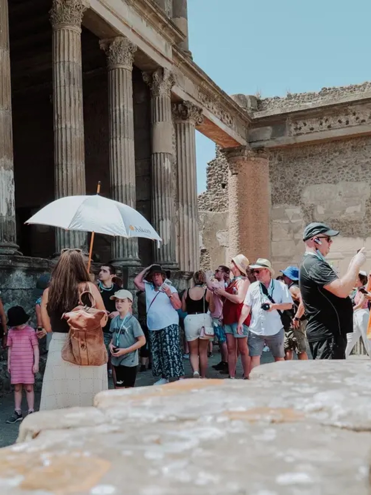 Tourists with umbrellas and cameras at ancient ruins with stone columns.