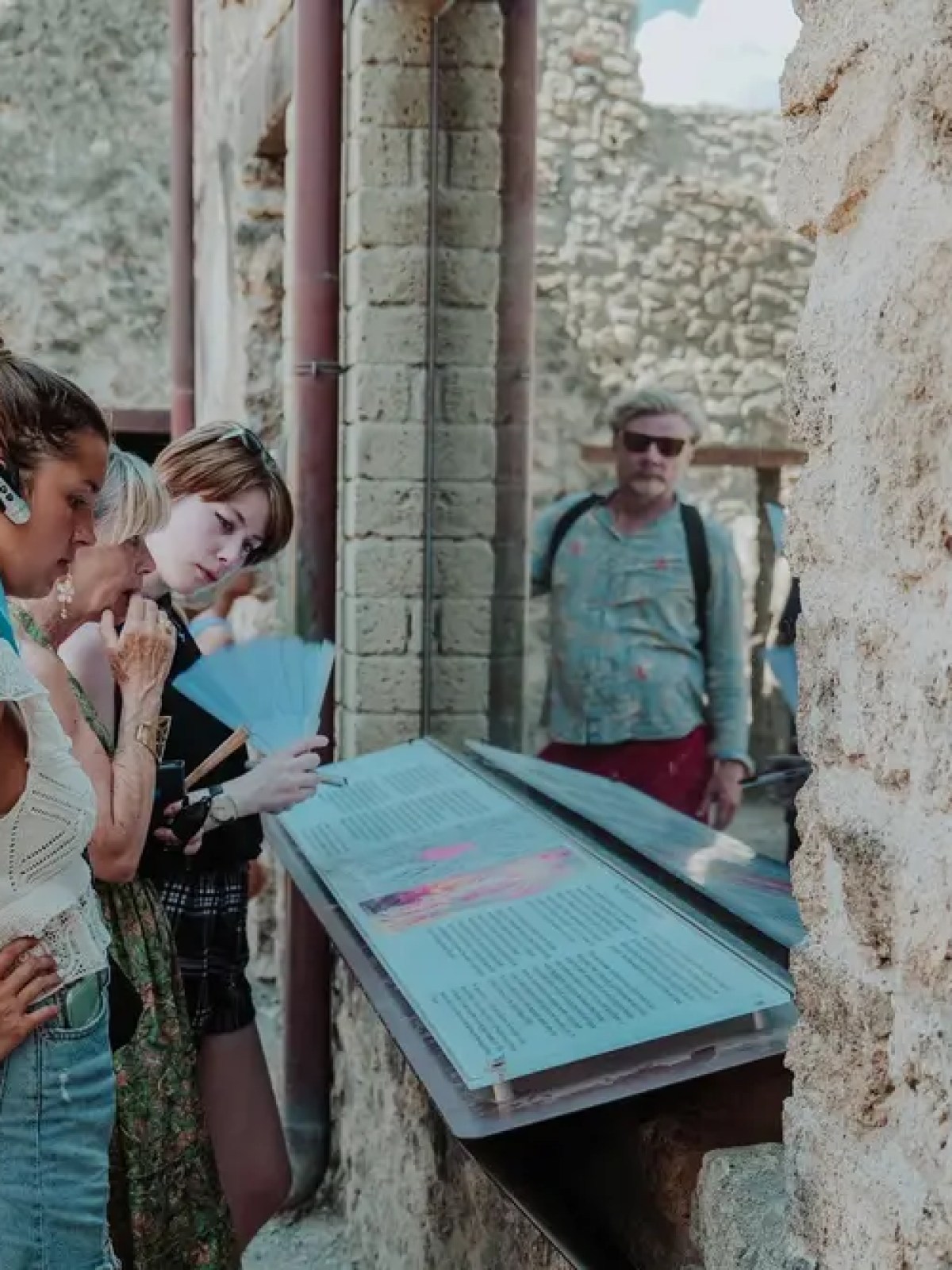 People reading information plaques at an ancient stone ruins site, with focus on a woman in a white top.
