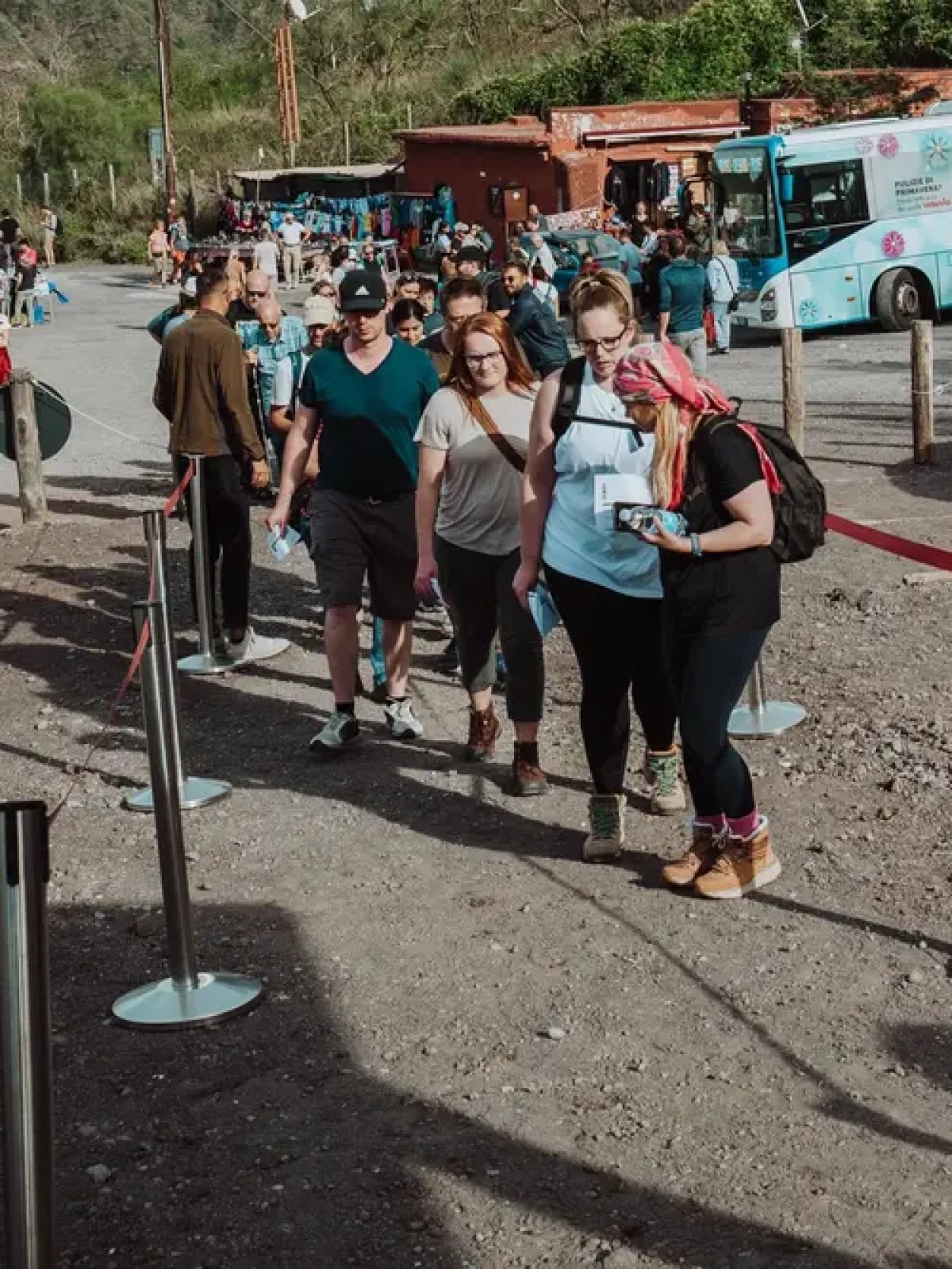 People in line near parked cars and a bus, outdoor setting with trees.