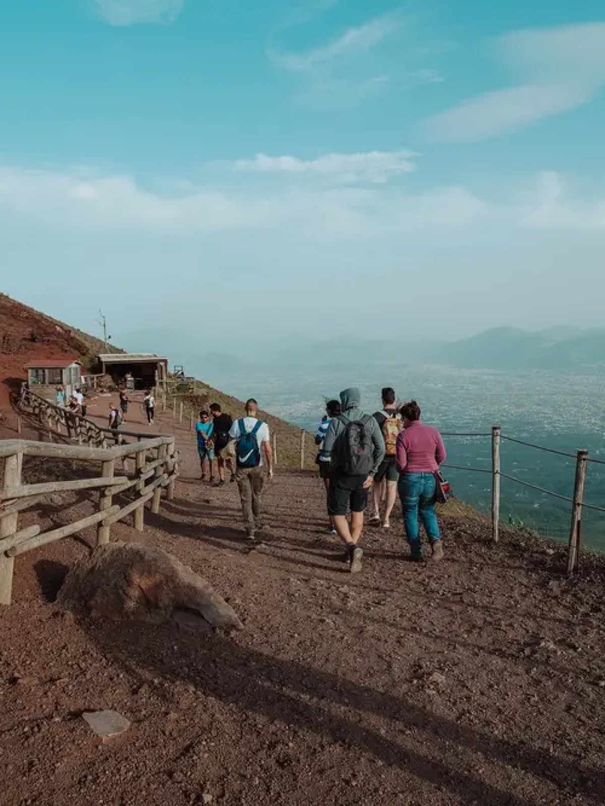 Hikers on a trail with a wooden fence and mountain view under a blue sky.