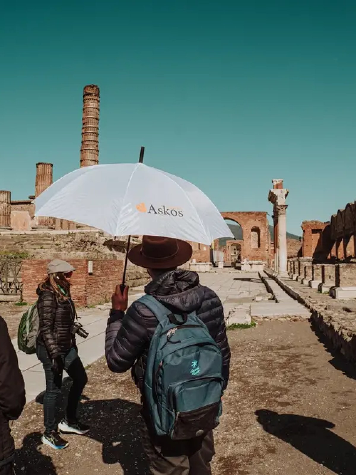 Group of tourists in ruins, one holding a white umbrella with 'Askos' logo.