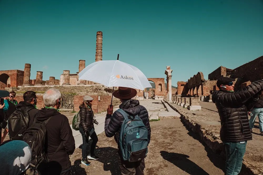 Group of tourists in ruins, one holding a white umbrella with 'Askos' logo.