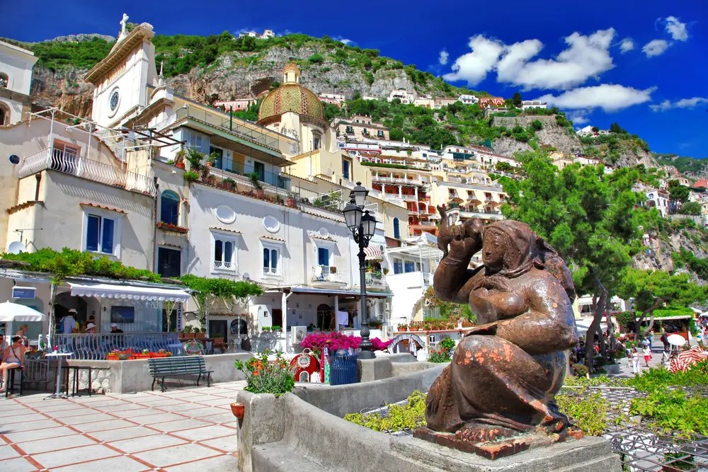 Statue in Positano plaza with colorful buildings and mountain backdrop under blue sky.