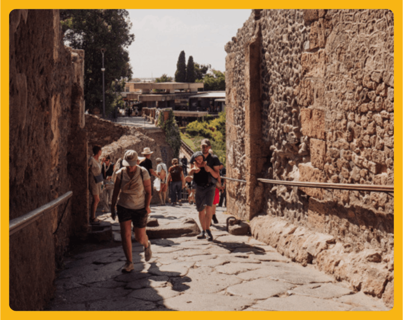 People walking on a stone path between ancient stone walls, surrounded by greenery.
