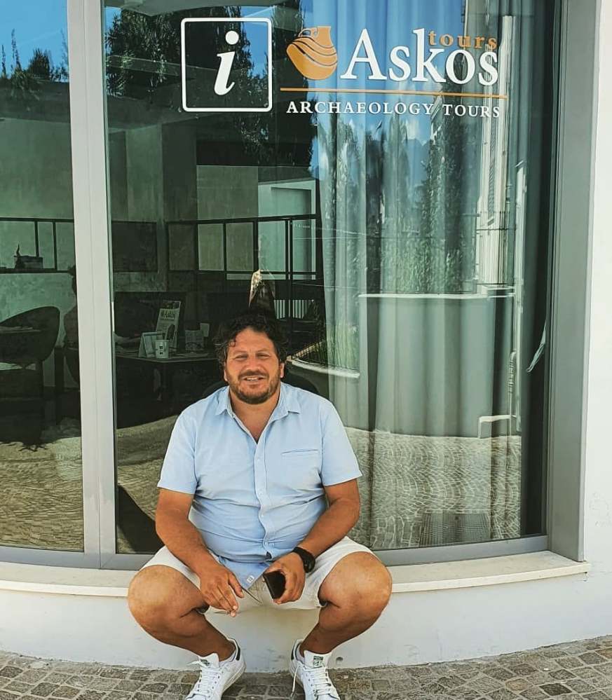 Man sitting in front of a glass window with 'Askos Archaeology Tours' logo.